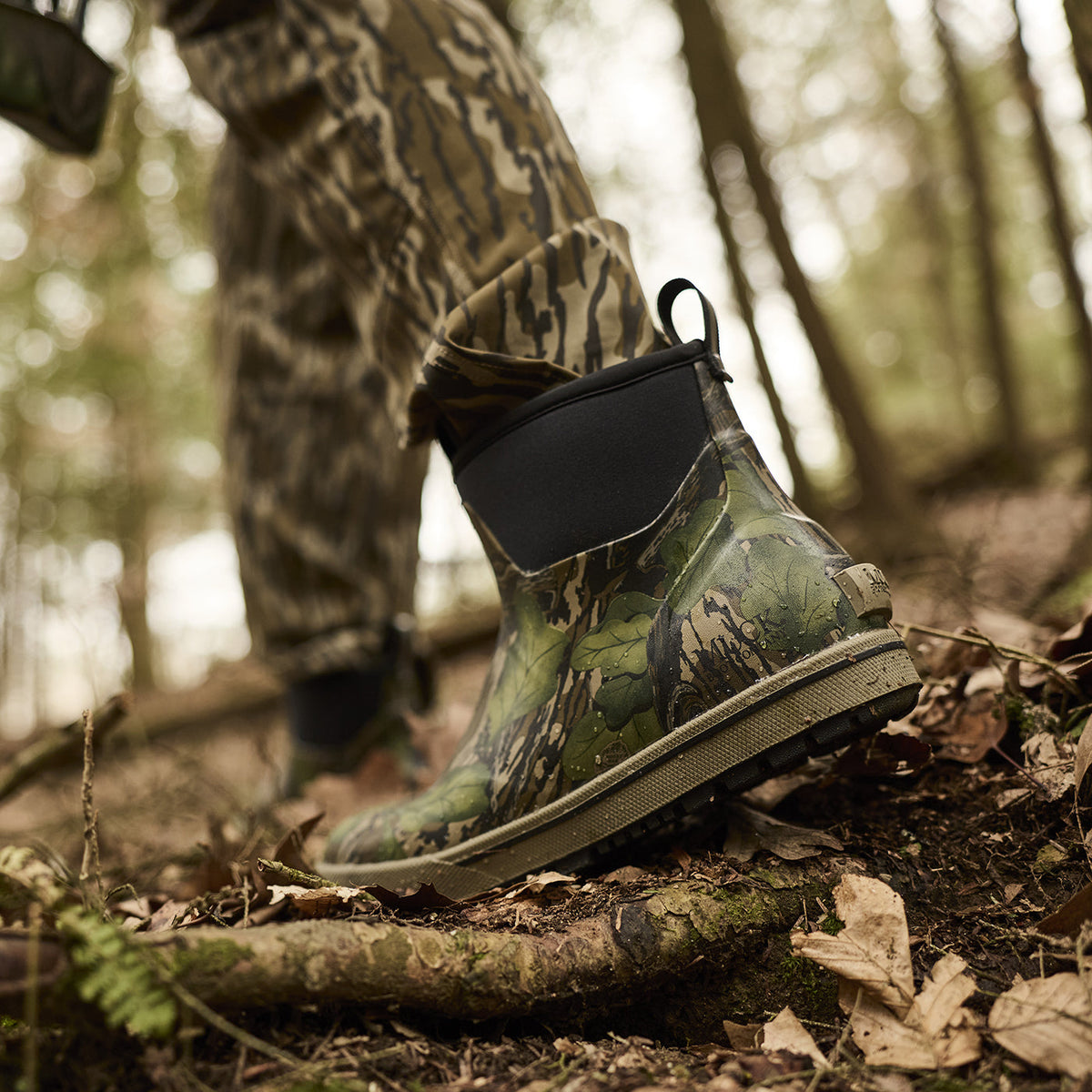 Camouflage boot on a forest floor with a person in camouflage clothing in the background.