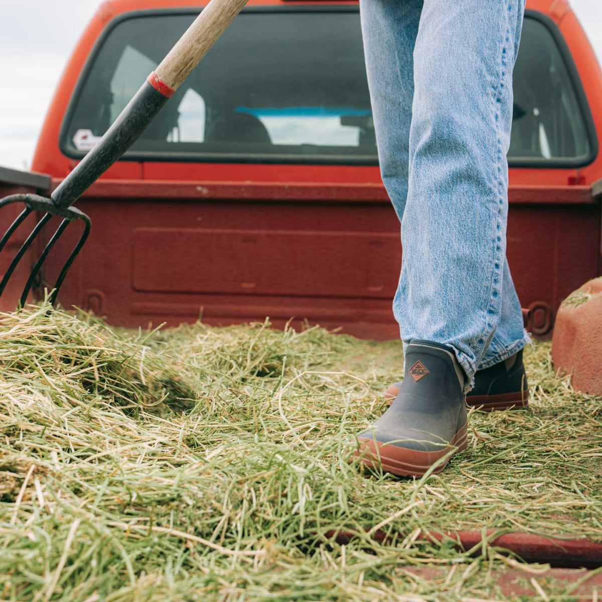 A person standing in a bed of hay next to a red pickup truck, wearing blue jeans and dark rubber boots, with a pitchfork visible in the foreground.