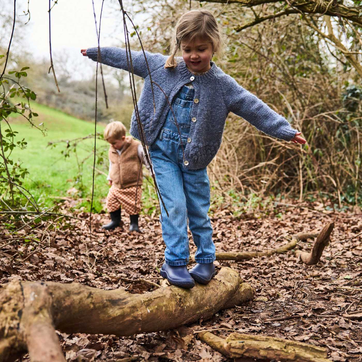 Two children playing in a forest, with one child balancing on a log.