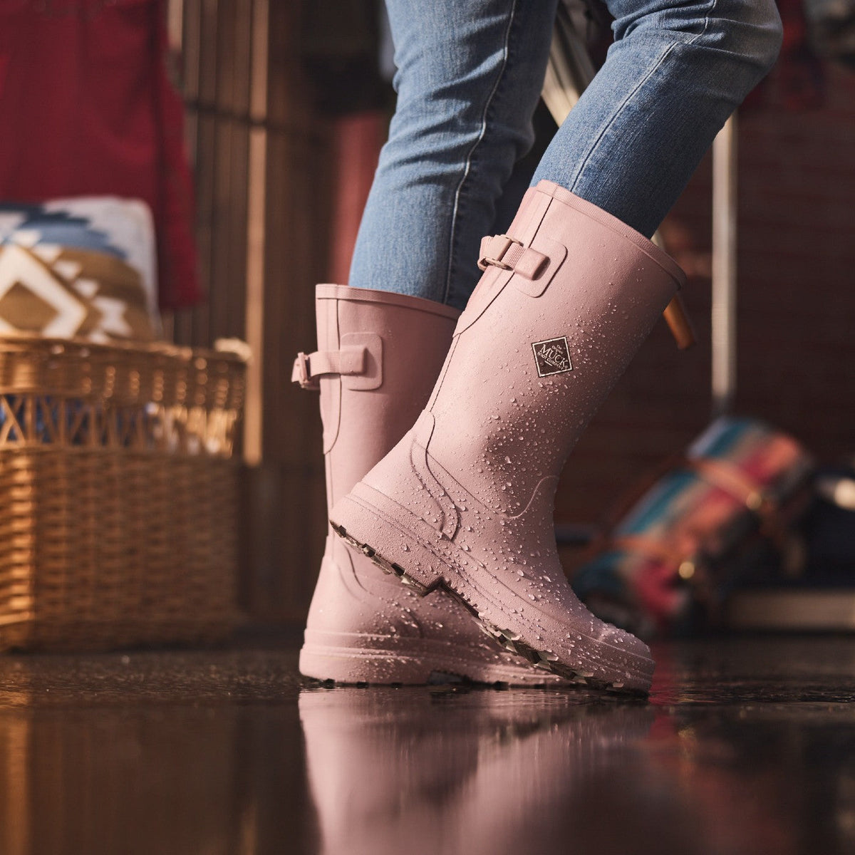 Pink rubber boots worn with blue jeans on a reflective surface, with a blurred indoor background.