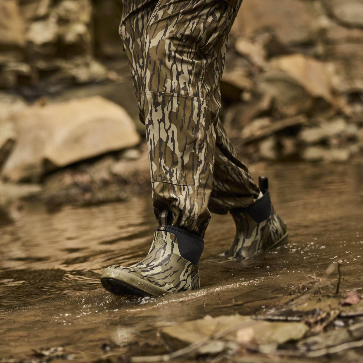Person wearing camouflage pants and Xplorer Short Waterproof Rubber boots in Mossy Oak Original Bottomland walking through water with rocks in the background